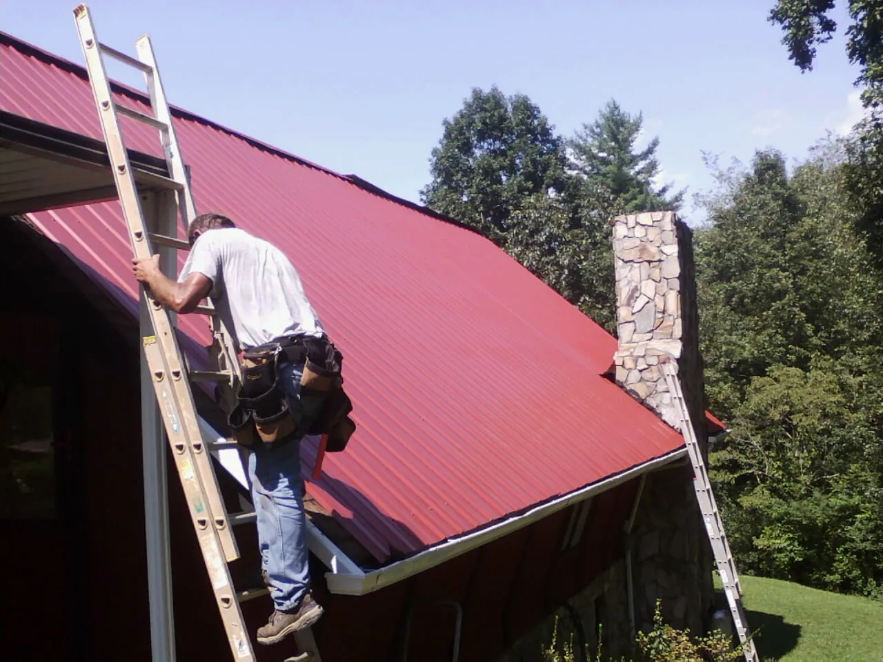 Roofer on a ladder beside a red metal roof and stone chimney — Soffit & Fascia Repair by Roofing Brothers in La Ward